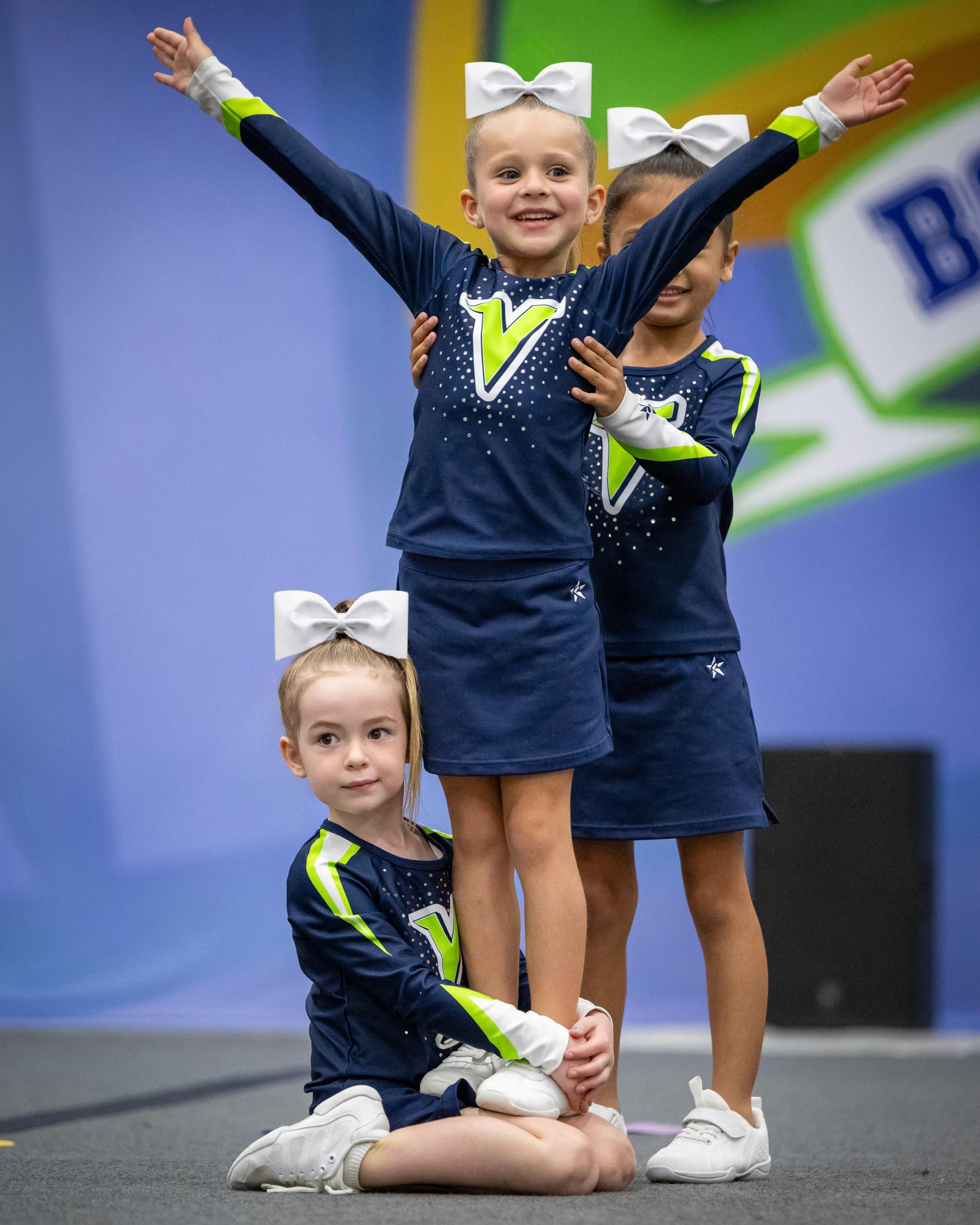 Young gymnasts in matching navy and lime uniforms with white bows perform a cheer pose on a blue backdrop; one child kneels while others stand with arms raised in triumph.