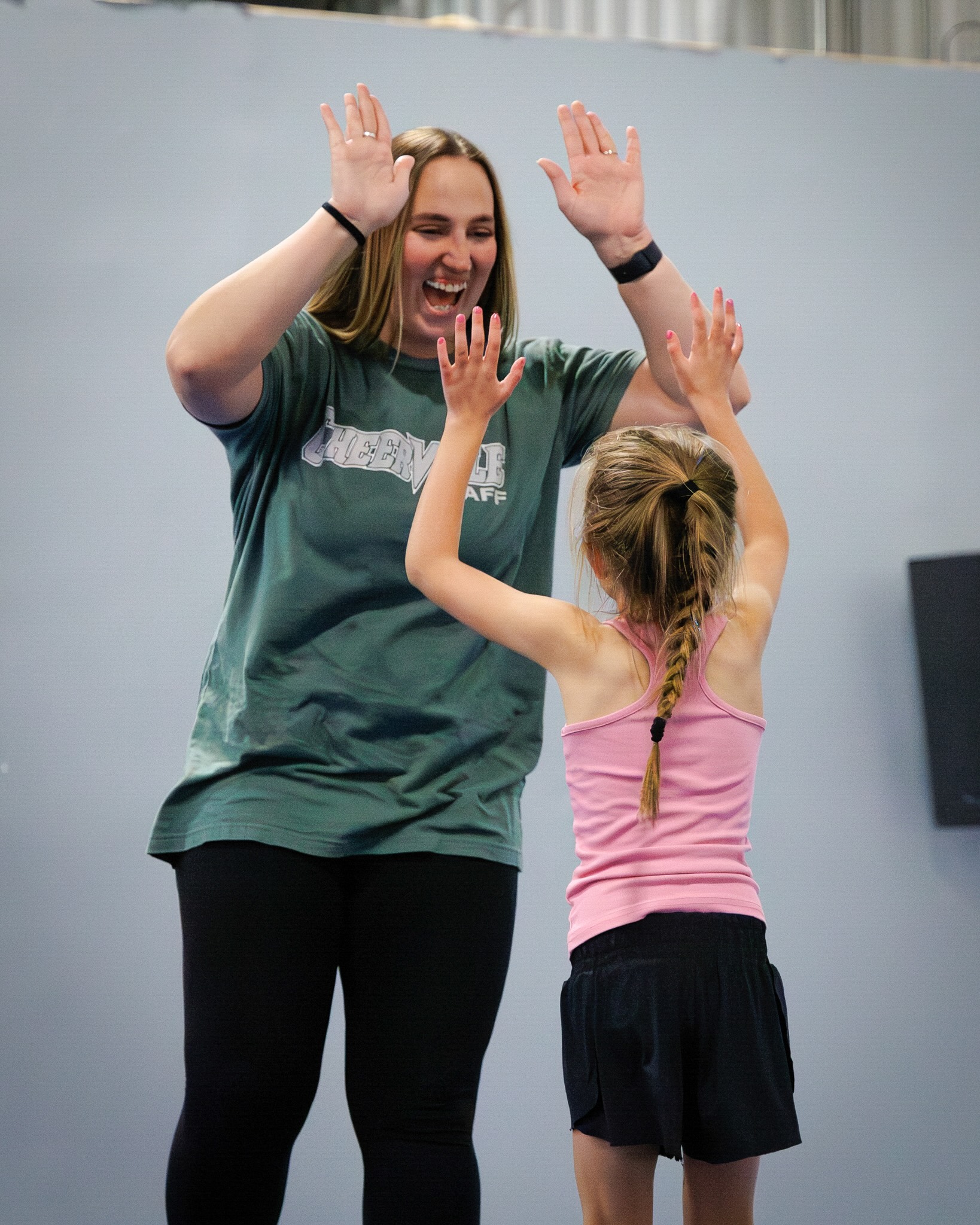Two people high-fiving indoors: a smiling woman in a green shirt reaching up with both hands toward a girl in a pink tank top who returns the high-five with pink painted nails.
