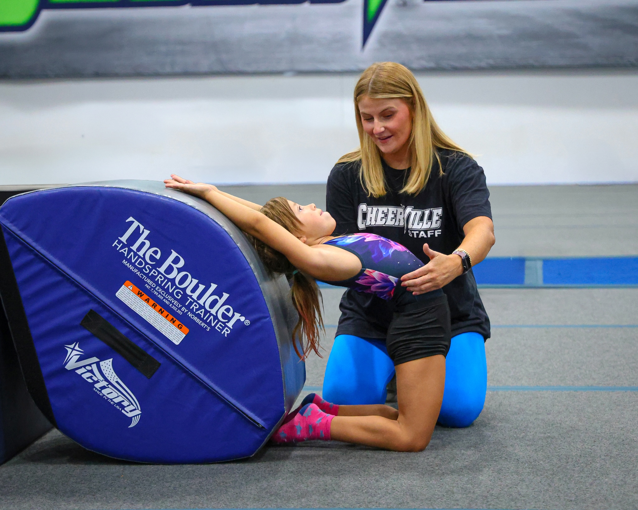 Female coach in a black shirt assists a young gymnast lying on a blue padded wedge in a gym setting.